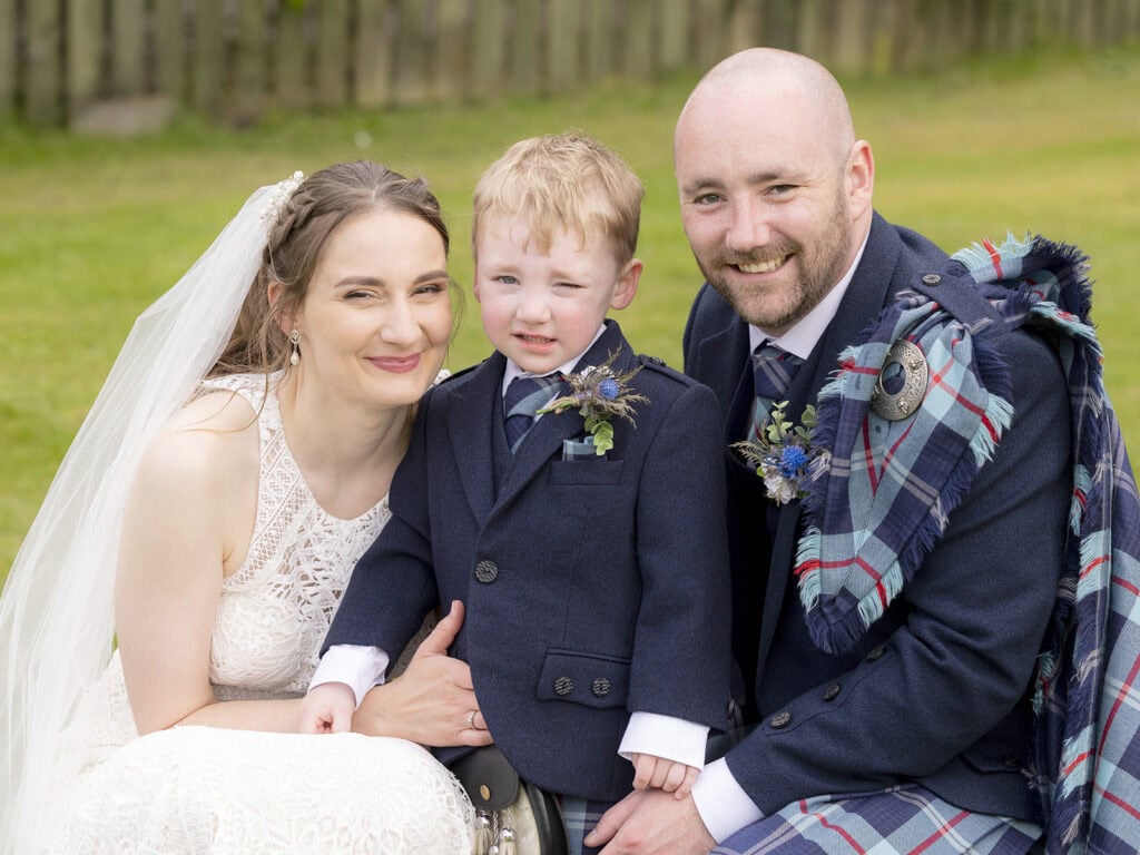 Bride and groom crouched beside their young son, all smiling in wedding outfits