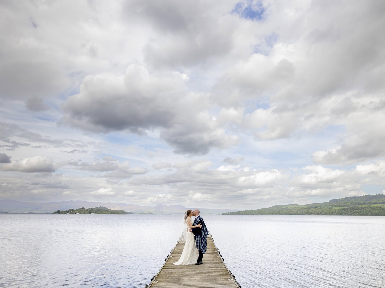 Bride and groom standing on a jetty at The Cruin, Loch Lomond, with dramatic sky and calm water behind them
